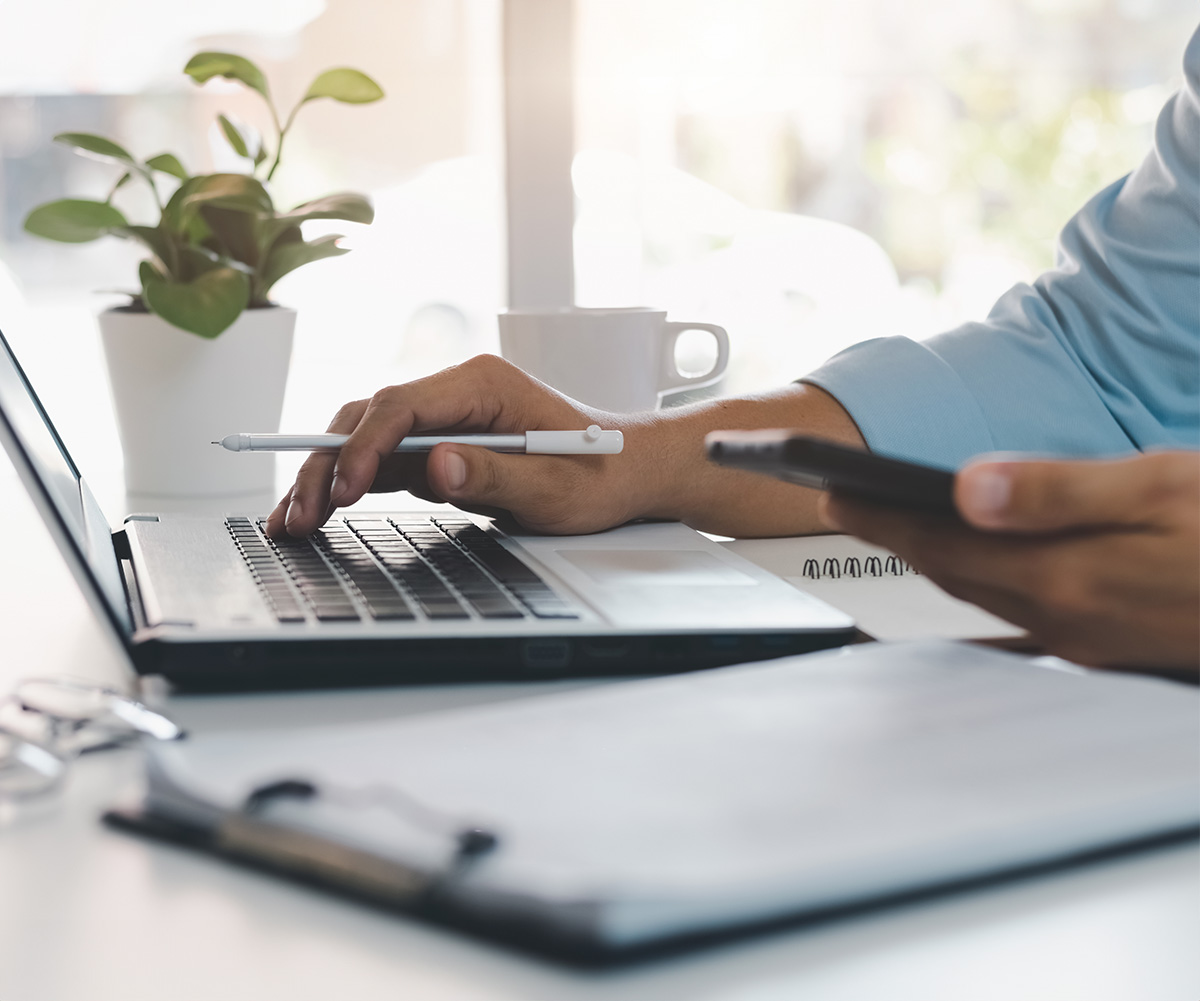 The image shows a person using a laptop at a desk with a smartphone nearby, surrounded by office items such as a pen, notebook, and potted plant, suggesting a professional work environment.