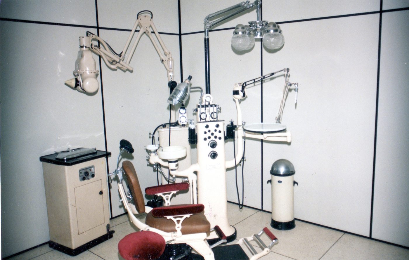 Mid-20th-century American dental operatory featuring multiple articulated arm-mounted instruments, an upholstered patient chair, and the cluster of lights and drills that defined a generation of dental visits.