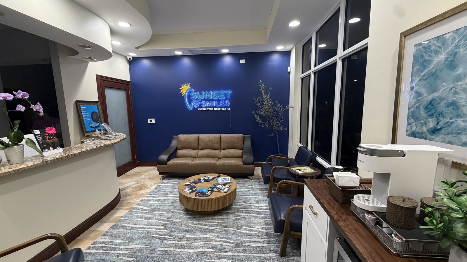 Newly remodeled Sunset Smiles Cosmetic Dentistry waiting area in Jupiter, FL, featuring the illuminated brand logo on the navy accent wall, leather seating and warm natural light