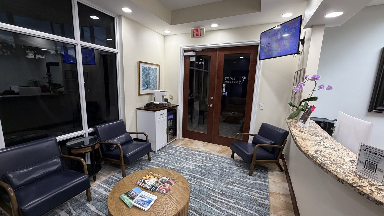 Patient waiting area at Sunset Smiles Cosmetic Dentistry Jupiter FL — modern lounge with tan leather sofa, navy chairs, floor-to-ceiling windows and complimentary Keurig coffee bar
