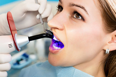 The image shows a woman receiving dental treatment, with a dental professional using a device to examine her teeth while she wears protective eyewear.