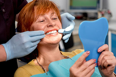 The image shows a woman with red hair sitting in a dental chair, holding a blue plastic model of a mouth, while a dental professional appears to be performing a procedure on her teeth using a drill.