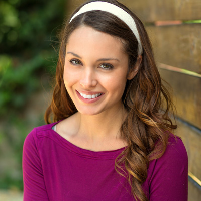 The image shows a woman with long brown hair smiling at the camera, wearing a purple top and a white headband, standing against a wooden fence.