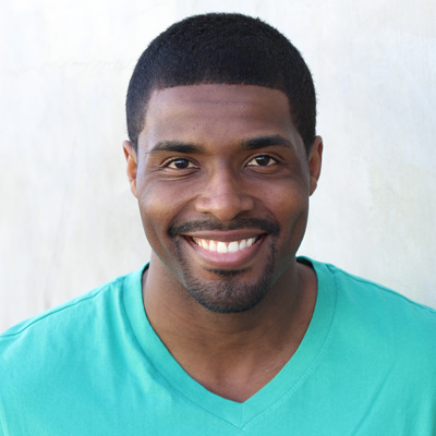 The image shows an elderly couple posing together with smiles, featuring a man on the left and a woman on the right, both dressed casually, with the man wearing a patterned shirt and the woman in a long-sleeved top. They are seated close to each other, embracing affectionately, set against a natural backdrop that includes trees and a clear sky.