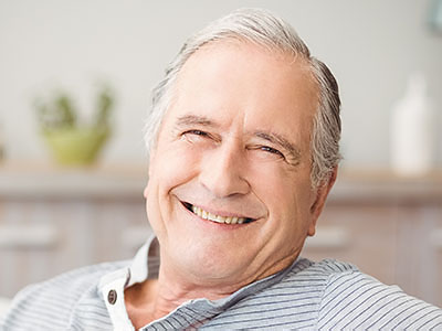 The image shows an elderly man with white hair, smiling and looking directly at the camera. He appears to be seated comfortably with his hands resting on a surface out of view.